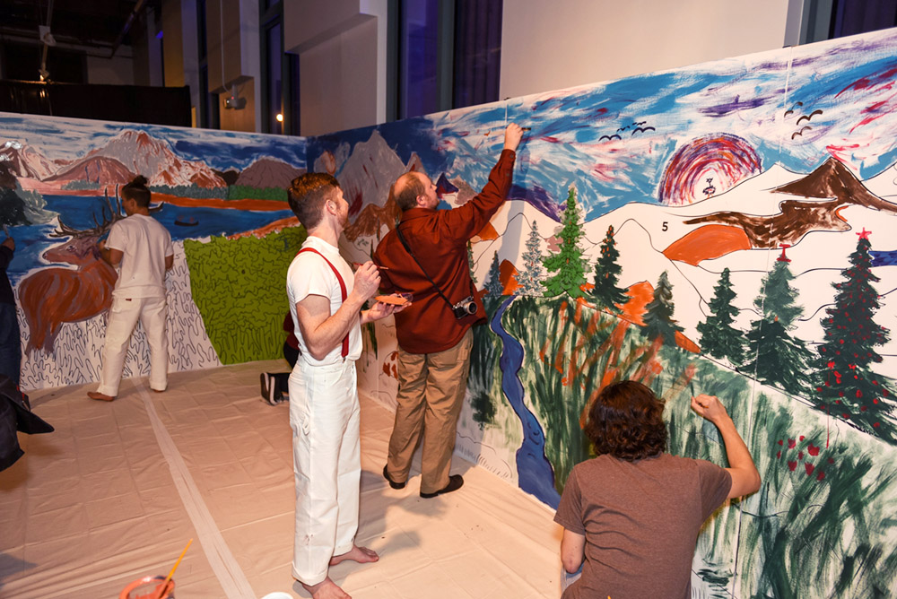 People help paint a large mural of mountains and trees.